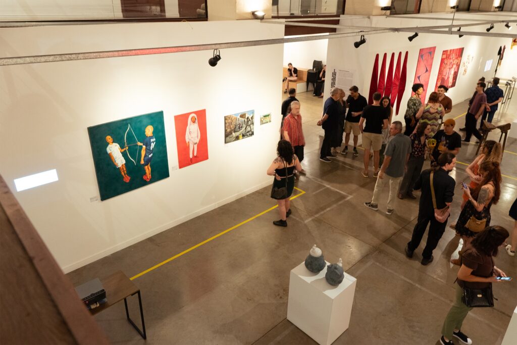 A imagem mostra um grupo de pessoas em uma galeria de arte, observando as obras expostas. Há várias pinturas na parede, algumas com figuras humanas e outras mais abstratas. No chão, há uma linha amarela que delimita o espaço da exposição. Algumas pessoas estão conversando, outras estão olhando atentamente para as obras. Há também algumas esculturas em pedestais. A iluminação é suave, criando uma atmosfera tranquila e contemplativa.