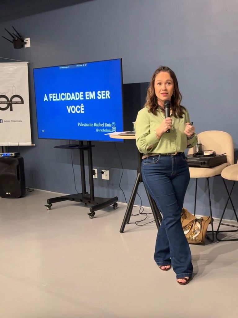 A mulher está em pé em frente a uma tela azul grande que exibe o título "A FELICIDADE EM SER VOCÊ" em letras brancas grandes. Abaixo do título, em letras menores, está escrito "Palestrante Rãchel Ruiz" e o @rachelruiz. A mulher está segurando um microfone em sua mão direita e parece estar falando. Ela está vestindo uma blusa verde clara e calças jeans escuras. A sala parece ser um local de apresentação, com um alto-falante à esquerda e cadeiras ao fundo.