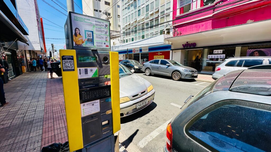 A imagem mostra uma cena de rua em uma cidade, provavelmente no Brasil, dado a presença de texto em português e o símbolo da moeda "R$". No primeiro plano, um parquímetro amarelo brilhante se destaca em um calçadão de ladrilhos. O parquímetro possui um display digital, um teclado e várias fendas para pagamento e emissão de tickets. Um código QR é visível no parquímetro, provavelmente para um aplicativo de estacionamento virtual. Acima do parquímetro, um grande painel publicitário exibe informações sobre a "Zona Azul Piracicaba", promovendo o uso de um aplicativo de estacionamento virtual e apresentando a imagem de uma mulher. A rua é ladeada por prédios que abrigam diversos comércios. Uma loja em destaque exibe o logo "vivo", uma empresa de telecomunicações, e uma placa anunciando "PS5 R$ 2.999". Mais adiante, outras lojas são visíveis, incluindo uma chamada "Drogal" e outra com "REMÉDIO DE".