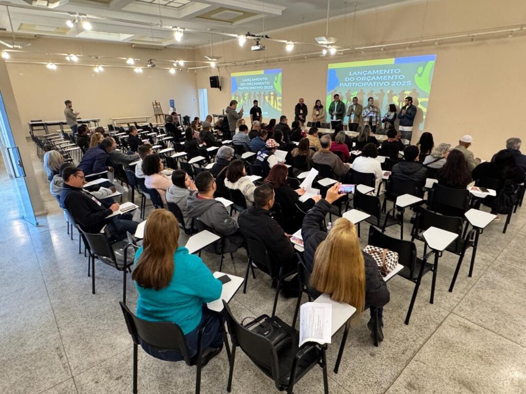 Na imagem, ocorre um evento em um auditório ou sala de aula, com várias pessoas sentadas em fileiras, acompanhando uma apresentação. Na frente, um grupo de palestrantes conduz a atividade, enquanto dois grandes telões exibem o título “Lançamento do Orçamento Participativo 2025”. O ambiente é moderno, bem iluminado e organizado, com participantes atentos, alguns fazendo anotações ou registrando o momento. A cena transmite a ideia de um encontro formal voltado à participação cidadã e ao planejamento financeiro público, reforçando o caráter democrático e coletivo da iniciativa.