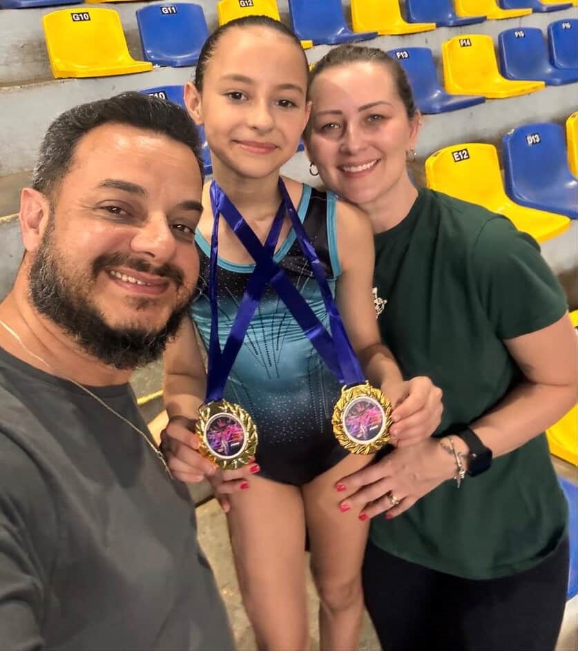 A imagem mostra uma família feliz posando para uma foto com uma jovem ginasta que acabou de ganhar medalhas. Na foto, vemos um homem sorridente com barba, uma menina com um uniforme de ginástica azul e preto e duas medalhas de ouro penduradas em seu pescoço, e uma mulher sorridente ao lado delas. Todos estão no que parece ser uma arena ou ginásio, com assentos azuis e amarelos ao fundo. A ginasta parece orgulhosa de suas conquistas, com um sorriso radiante. Os pais também expressam alegria e orgulho ao lado de sua filha. As medalhas douradas brilham, com fitas azuis penduradas, destacando o sucesso da jovem atleta. A atmosfera geral da imagem é de celebração e união familiar.