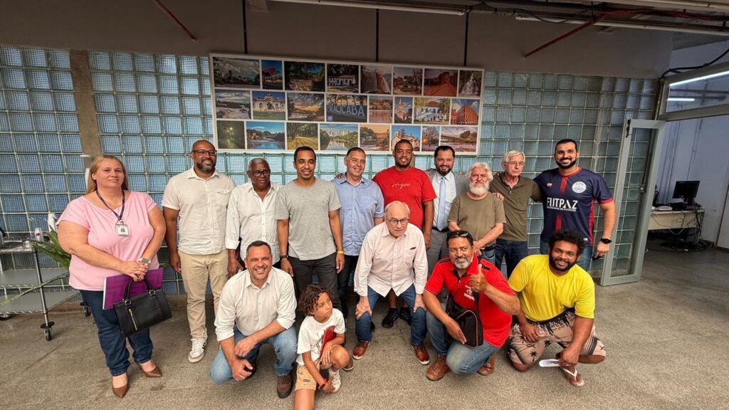 A fotografia mostra um grupo de 14 pessoas, 13 homens e 1 mulher, posando para uma foto em um ambiente interno, possivelmente um escritório ou corredor. Atrás deles, há uma parede decorada com uma colagem de várias fotografias de paisagens urbanas e naturais, com a palavra "PIRACICABA" em destaque em uma das imagens. A mulher, à esquerda, veste uma blusa rosa e jeans, e segura uma bolsa preta e alguns papéis. Os homens presentes exibem uma variedade de idades e etnias, vestindo roupas casuais e formais. Alguns estão em pé, enquanto outros estão agachados na frente do grupo. Um dos homens usa uma camiseta de futebol com a inscrição "FUTPAZ" e "ESCOLA DE FUTEBOL". A iluminação na cena é artificial e parece vir de cima.