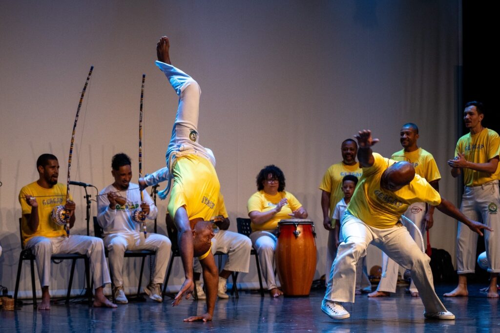 A imagem retrata um grupo de pessoas praticando capoeira em um palco. No centro, um homem executa um movimento acrobático de cabeça para baixo, vestindo uma camiseta amarela e calças brancas. Ao redor dele, outros participantes tocam instrumentos musicais típicos da capoeira, como o berimbau e o atabaque, enquanto outros observam ou se preparam para dançar. A iluminação do palco destaca os artistas, criando um ambiente de performance e celebração da cultura afro-brasileira.