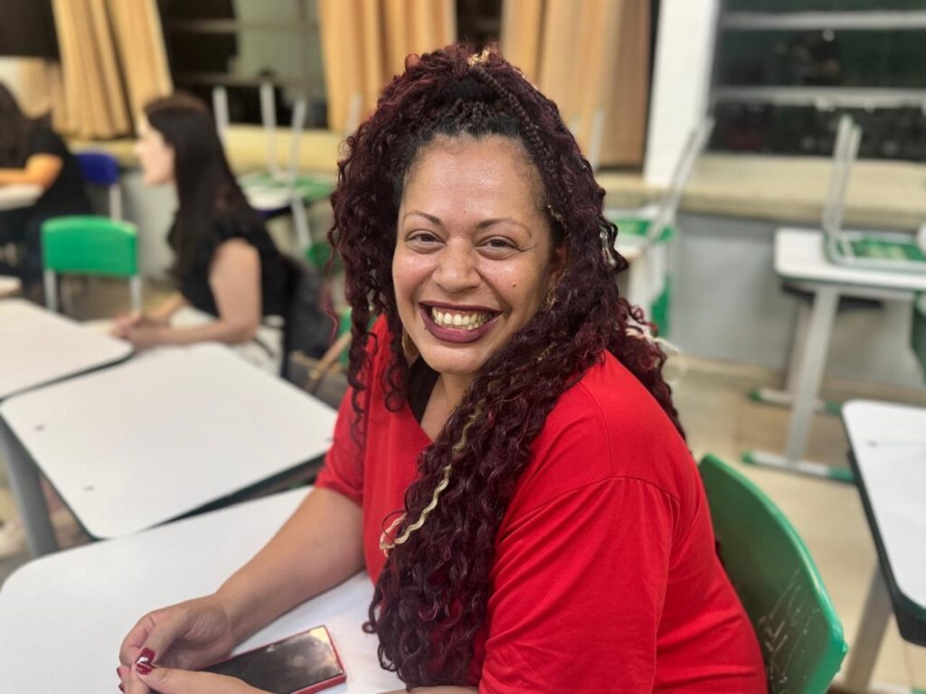 A mulher sorri para a câmera, sentada em uma sala de aula com carteiras e cadeiras. Ela tem cabelos longos e ondulados de cor vinho, com algumas tranças. Ela usa uma blusa vermelha e tem unhas pintadas de vermelho escuro. Um celular vermelho está sobre a carteira à sua frente. Ao fundo, outras pessoas estão sentadas em carteiras, e algumas cadeiras estão empilhadas. As cortinas ao fundo são de cor bege.