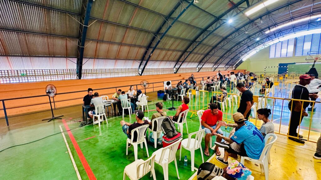 A imagem mostra um grande salão com teto arqueado, que parece ser uma quadra esportiva, com o chão pintado de verde. Várias pessoas estão reunidas em grupos, sentadas em cadeiras brancas ao redor de mesas. Há mesas de jogo ou de trabalho espalhadas pelo local. No lado esquerdo da imagem, há um ventilador grande em pé. Mais ao fundo, observa-se uma arquibancada laranja e uma grade de proteção. Perto da entrada, algumas pessoas estão em pé, conversando. No centro e à direita, há mais mesas com pessoas sentadas, algumas interagindo e outras parecendo esperar. Há também cercas amarelas delimitando algumas áreas. A iluminação vem de cima, através do teto. Algumas pessoas estão usando máscaras. A cena geral sugere um evento, uma reunião, ou talvez algum tipo de serviço sendo prestado, com as pessoas organizadas em torno de mesas para diferentes atividades.