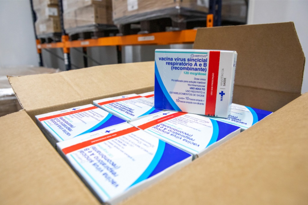 A imagem mostra uma caixa de papelão aberta, revelando várias caixas de um medicamento. Uma das caixas está em destaque, mostrando claramente o rótulo com o nome "vacina vírus sincicial respiratório A e B (recombinante)". A embalagem indica que a dose é de 120 mcg, é uma dose única, para solução injetável, administrada por via intramuscular, destinada a uso adulto e restrita a estabelecimentos de saúde. A caixa contém 10 frascos-ampola com diluente. A embalagem também exibe o logo do Ministério da Saúde do Brasil e a sigla SUS. Ao fundo, embaçadas, podem ser vistas prateleiras de um armazém com caixas de outros produtos, sugerindo que se trata de um local de armazenamento ou distribuição.