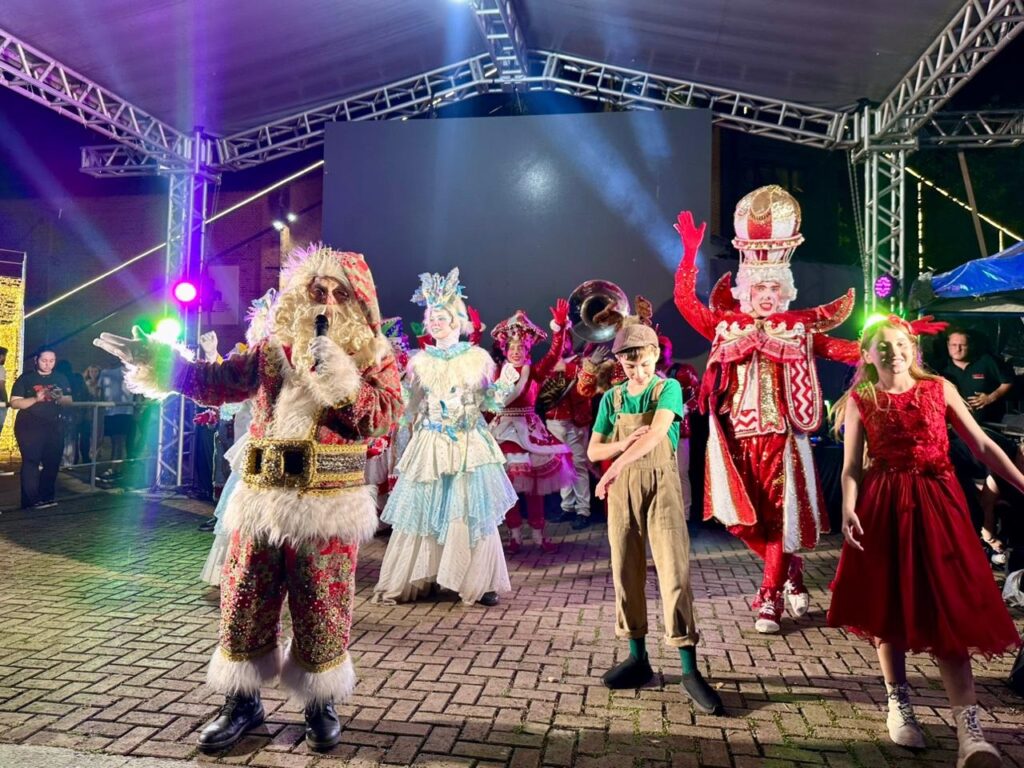 A imagem mostra um grupo de artistas fantasiados em um palco iluminado. No centro, um homem vestido como Papai Noel, com um traje vermelho brilhante adornado com pelos brancos e detalhes dourados, segura um microfone e gesticula com a mão. Ao seu lado, há uma figura alta com uma fantasia vermelha e branca, usando um chapéu elaborado, e uma menina com um vestido vermelho. Atrás deles, outros artistas em trajes festivos, incluindo um músico com um instrumento de sopro, completam a cena. O palco é decorado com luzes coloridas e uma tela grande ao fundo. O chão é pavimentado com pedras.