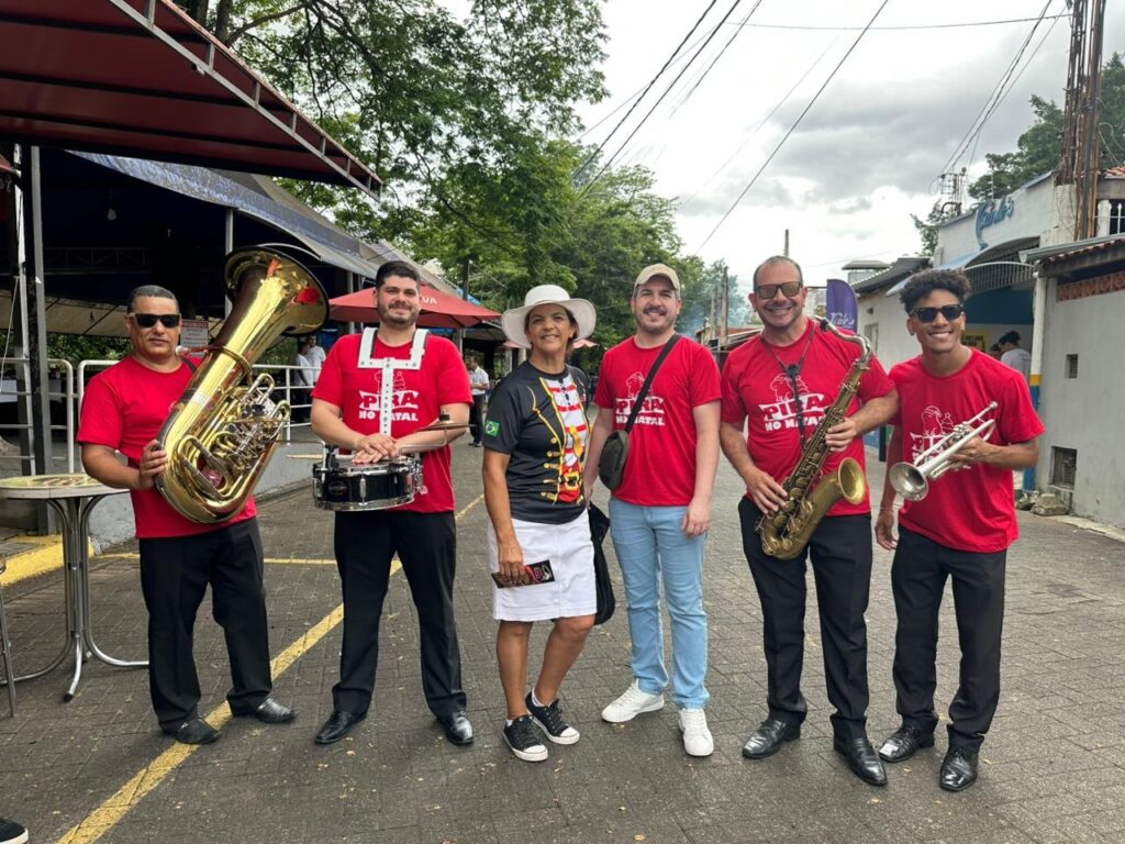 A imagem mostra um grupo de seis pessoas, cinco homens e uma mulher, posando para uma foto ao ar livre. Todos, exceto a mulher no centro, usam camisetas vermelhas com uma estampa que parece ser um logo. A mulher usa uma camiseta preta com detalhes em amarelo e vermelho, e uma saia branca. Os instrumentos musicais presentes são um tuba, uma caixa, um saxofone e uma trombeta. A pessoa mais à esquerda segura a tuba, a segunda pessoa da esquerda segura a caixa, a quarta pessoa da esquerda segura o saxofone e a pessoa mais à direita segura a trombeta. O grupo parece estar em uma rua, com construções ao fundo e árvores em ambos os lados. O céu está nublado. A iluminação sugere que a foto foi tirada durante o dia. A atmosfera geral é informal e festiva, possivelmente indicando uma apresentação musical ou um evento comunitário.