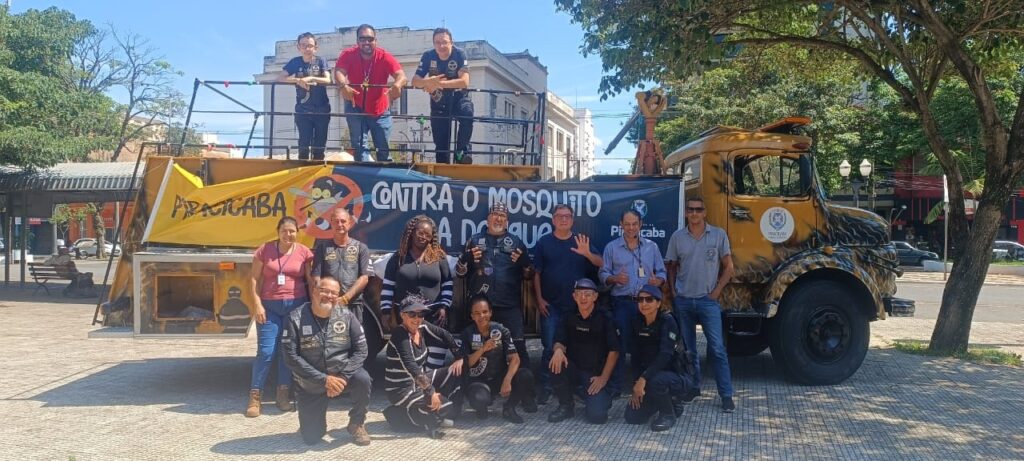 A imagem mostra um grupo de pessoas em frente a um caminhão decorado. O caminhão tem uma pintura amarela com detalhes pretos que lembram chamas ou um padrão de animal. Na lateral do caminhão, há um banner amarelo com a inscrição "Piracicaba" e outro banner escuro com a frase "Contra o Mosquito da Dengue". As pessoas estão posicionadas em duas fileiras. Na fileira da frente, algumas pessoas estão agachadas ou ajoelhadas, enquanto outras estão em pé. Muitas delas usam coletes pretos com emblemas, sugerindo que fazem parte de alguma organização ou grupo. Na fileira de trás, algumas pessoas estão em pé ao lado do caminhão, e outras estão em uma plataforma elevada no veículo. O cenário parece ser uma área urbana com prédios ao fundo e árvores. O céu está claro, indicando um dia ensolarado.
