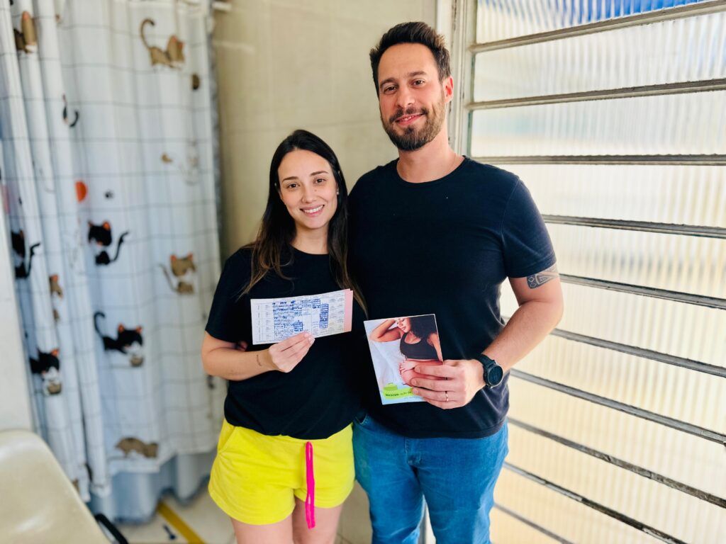 A imagem mostra um casal sorrindo em frente a uma janela com persianas translúcidas. A mulher, à esquerda, veste uma camiseta preta e shorts amarelos vibrantes com um cordão rosa. Ela segura um pequeno livreto ou documento com várias linhas de texto azul e rosa. Ao lado dela, o homem, à direita, veste uma camiseta preta e jeans azuis. Ele exibe uma tatuagem no braço esquerdo e segura um cartão ou folheto com a imagem de uma mulher grávida na capa. O fundo atrás da mulher é uma cortina branca com um padrão lúdico de gatos pretos e marrons. A iluminação geral sugere que a foto foi tirada durante o dia, com luz natural entrando pela janela.