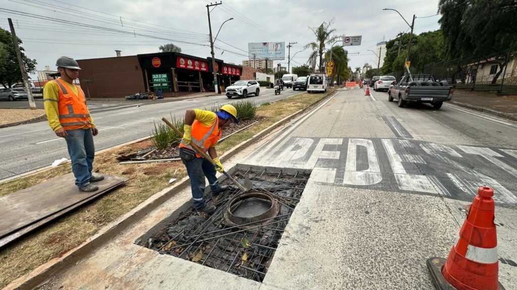 A imagem mostra dois trabalhadores da construção civil em uma área urbana, com foco em uma vala aberta na calçada que parece ser para manutenção de um bueiro. Um trabalhador, vestindo um colete laranja de alta visibilidade sobre uma camisa amarela e calças jeans, está ajoelhado dentro da vala, usando luvas amarelas e um capacete amarelo. Ele está usando uma pá para trabalhar no bueiro, que está exposto com vergalhões de metal e uma cobertura preta por baixo. O outro trabalhador, vestindo um colete laranja semelhante sobre uma camisa amarela e calças jeans, está em pé na calçada ao lado da vala, usando um capacete cinza. Ele está parado sobre algumas chapas de metal. A vala está localizada ao lado de uma rua movimentada com tráfego de veículos, incluindo carros, vans e uma caminhonete. Há placas de trânsito e semáforos visíveis ao fundo, bem como edifícios comerciais e residenciais. Uma laranja de trânsito está posicionada perto da vala para sinalizar a área de trabalho. As palavras "PEDEST" estão pintadas na estrada em frente à vala, indicando uma área de pedestres ou uma faixa de pedestres. O céu está nublado, sugerindo um dia com pouca luz.