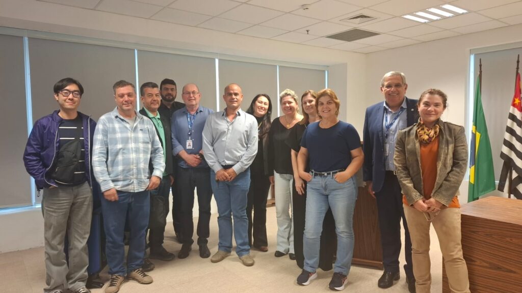 A foto mostra um grupo de treze pessoas reunidas em um ambiente interno bem iluminado, com teto de placas e grandes janelas cobertas por persianas. Elas estão alinhadas lado a lado, posando para o registro, vestindo roupas que variam entre o casual e o social. Ao fundo, destacam-se duas bandeiras: a do Brasil e a do estado de São Paulo, sugerindo que o encontro ocorreu em um espaço institucional ou governamental. A cena transmite a ideia de um momento oficial ou profissional, marcado pela formalidade e pela representatividade do grupo presente.
