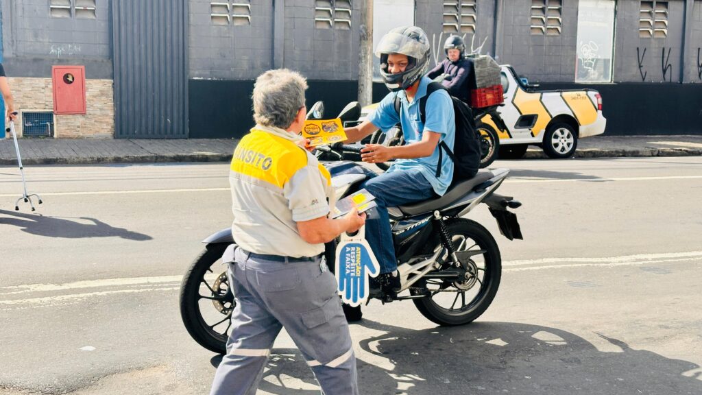 Na imagem, uma agente de trânsito com uniforme amarelo e cinza, ostentando a palavra "TRÂNSITO" nas costas, aborda um motociclista. A agente segura um panfleto e um objeto em formato de luva azul com a inscrição "ATENÇÃO! RESPEITE A FAIXA". O motociclista, vestindo capacete e camiseta azul, está parado em sua moto e parece receber o material informativo. Ao fundo, um veículo utilitário branco com detalhes amarelos e a placa "3407-1111" está estacionado, com outra pessoa usando capacete no banco do passageiro. Há também uma pessoa com muletas em primeiro plano, à esquerda, e uma parede de tijolos e concreto ao fundo, sugerindo um ambiente urbano. A luz do sol cria sombras nítidas no asfalto.