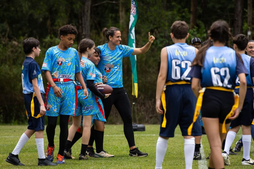 A imagem mostra um grupo de crianças e uma mulher em um campo de futebol gramado. A maioria das crianças está vestindo uniformes azuis claros com desenhos e o texto "Canera" e "Delta". As crianças em primeiro plano parecem estar participando de um jogo de flag football, pois algumas usam faixas coloridas presas à cintura. A mulher, que está no centro da imagem, veste uma camiseta azul semelhante e calças pretas. Ela está segurando uma bola de futebol americano e parece estar instruindo ou motivando as crianças, com o dedo indicador levantado. Atrás dela, há uma bandeira verde e branca com o logotipo da NFL. Outras crianças estão em segundo plano, algumas vestindo uniformes azuis com o número "62" nas costas. O ambiente sugere um treinamento ou jogo ao ar livre, com árvores visíveis ao fundo. A iluminação indica que é dia.