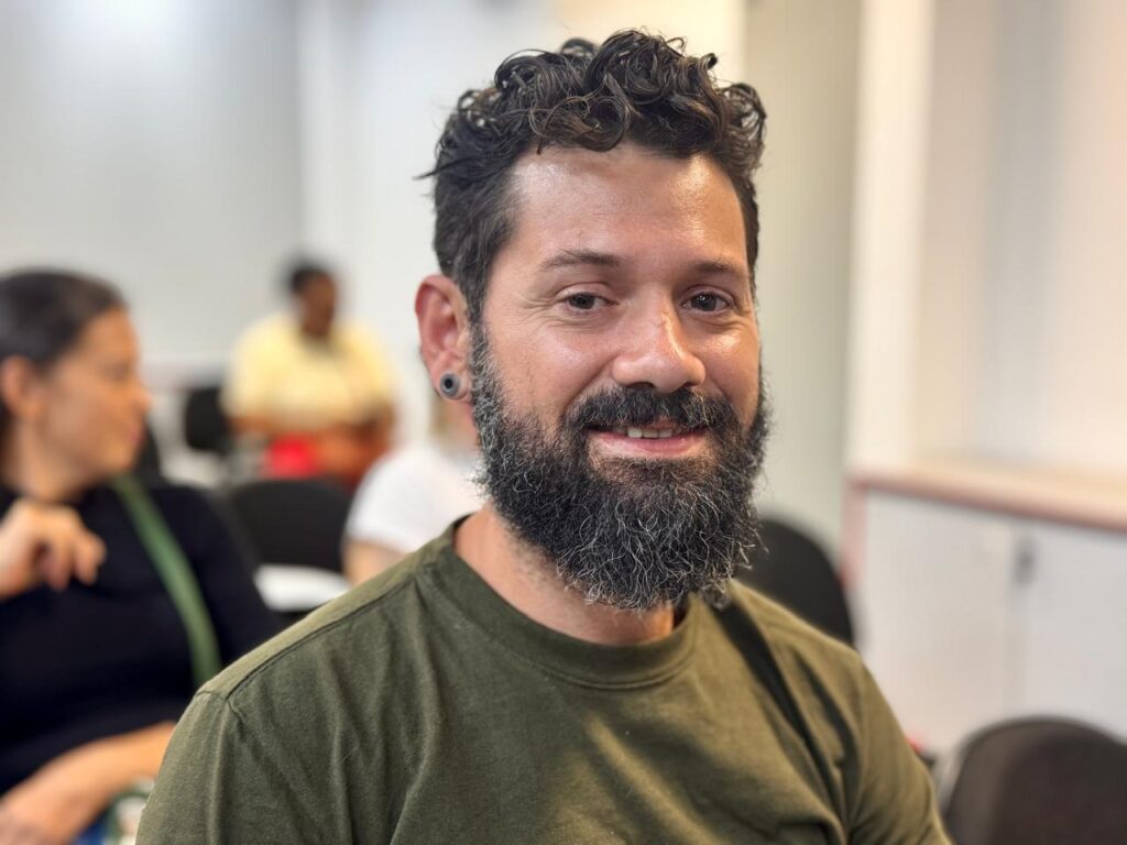 A imagem mostra um close-up de um homem com barba e cabelo encaracolado, vestindo uma camiseta verde. Ele está sorrindo e olhando para a câmera. Ao fundo, desfocadas, vêem-se outras pessoas sentadas em cadeiras, possivelmente em uma sala de aula ou reunião. O ambiente parece ser um local interno, com paredes claras e algumas estruturas de móveis ao fundo. A iluminação é suave e natural.