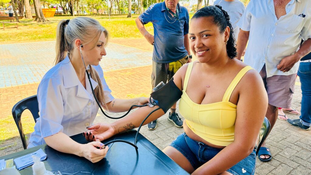 A imagem mostra uma profissional de saúde, com jaleco branco e cabelos presos em um rabo de cavalo, sentada e medindo a pressão de uma mulher sorridente. A mulher, que usa um top amarelo e shorts jeans, está sentada em uma cadeira preta e olha para a câmera com um sorriso. Ao fundo, outras pessoas estão em pé, em um ambiente externo com árvores e um piso de paralelepípedos. Há uma mesa à frente da profissional de saúde com alguns papéis e um frasco de álcool em gel.