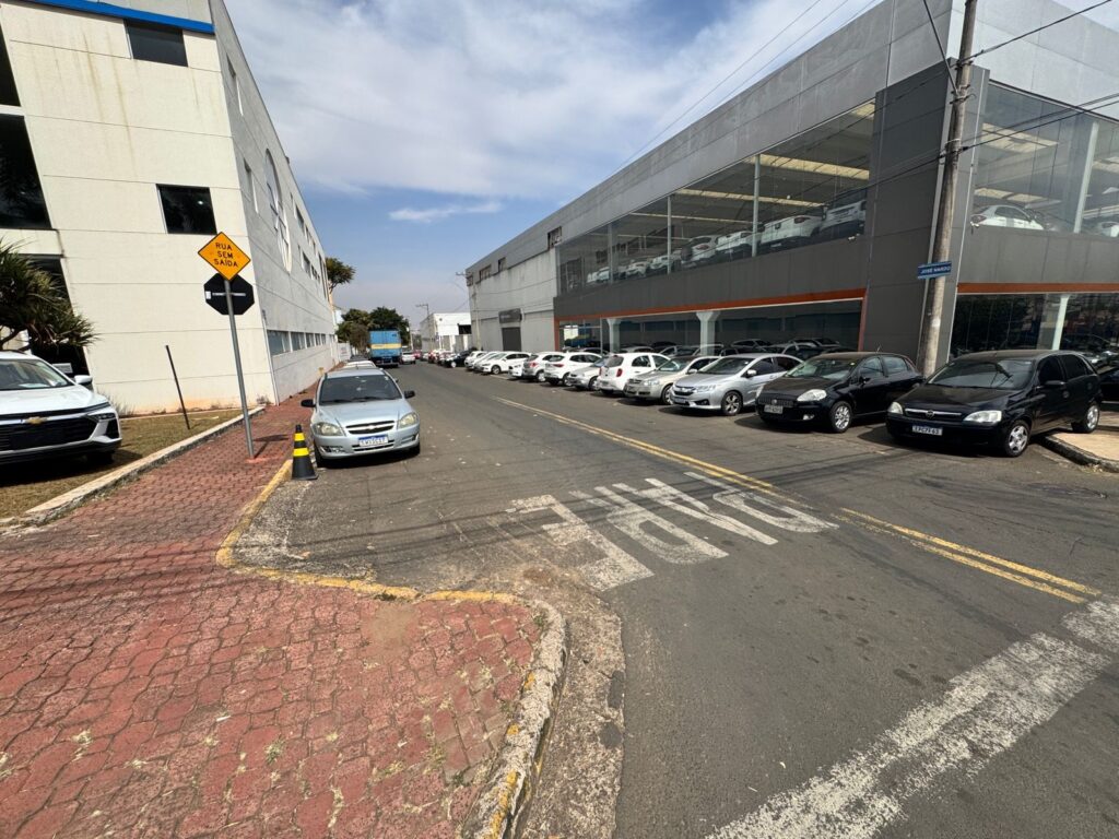 A imagem mostra uma rua com vários carros estacionados em frente a uma concessionária de automóveis. A concessionária tem uma fachada moderna com grandes janelas de vidro, permitindo ver os carros expostos no interior. Do lado esquerdo da rua, há um prédio de escritórios com uma arquitetura angular e um sinal de "Rua sem saída". Vários carros estão estacionados ao longo da rua, tanto na lateral da concessionária quanto do outro lado. Há também um caminhão azul ao fundo. O asfalto da rua tem marcações de faixa e a palavra "PARE" pintada em branco. O céu está parcialmente nublado, indicando um dia claro.
