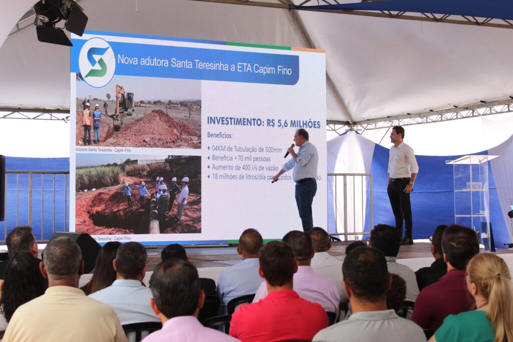 A imagem mostra uma apresentação sobre uma nova adutora em um evento ao ar livre, possivelmente uma inauguração ou anúncio. No centro da imagem, um grande telão exibe informações sobre a "Nova adutora Santa Teresinha a ETA Capim Fino". O telão contém imagens do projeto, detalhes do investimento e os benefícios esperados. Um homem de camisa azul e calça jeans está de pé em frente ao telão, falando em um microfone, aparentemente o orador principal. À direita do orador, outro homem de camisa branca e calça preta está em pé, observando o telão ou a audiência. Na frente do telão e dos palestrantes, uma audiência está sentada em cadeiras, voltada para a apresentação. A maioria das pessoas na audiência está de costas para a câmera, com suas cabeças visíveis. O ambiente é coberto por uma estrutura de tenda branca, com detalhes em azul. Há também uma grade metálica e um púlpito transparente visíveis ao fundo. A iluminação sugere que o evento está ocorrendo durante o dia.
