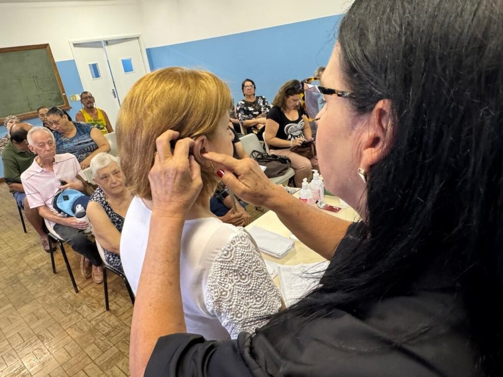 A imagem mostra um grupo de pessoas, a maioria idosos, sentados em cadeiras enfileiradas em uma sala. No centro, uma mulher de cabelos ruivos está sendo atendida por outra mulher, que usa óculos e tem cabelos pretos longos. A atendente parece estar examinando ou tratando o ouvido da mulher ruiva com um instrumento fino e um frasco pequeno na mão. As outras pessoas na sala observam a cena com atenção, algumas parecendo esperar sua vez. O ambiente parece ser um local público, possivelmente uma clínica ou centro comunitário, com uma lousa e portas ao fundo. Há uma atmosfera de cuidado e atenção médica sendo prestada aos idosos.