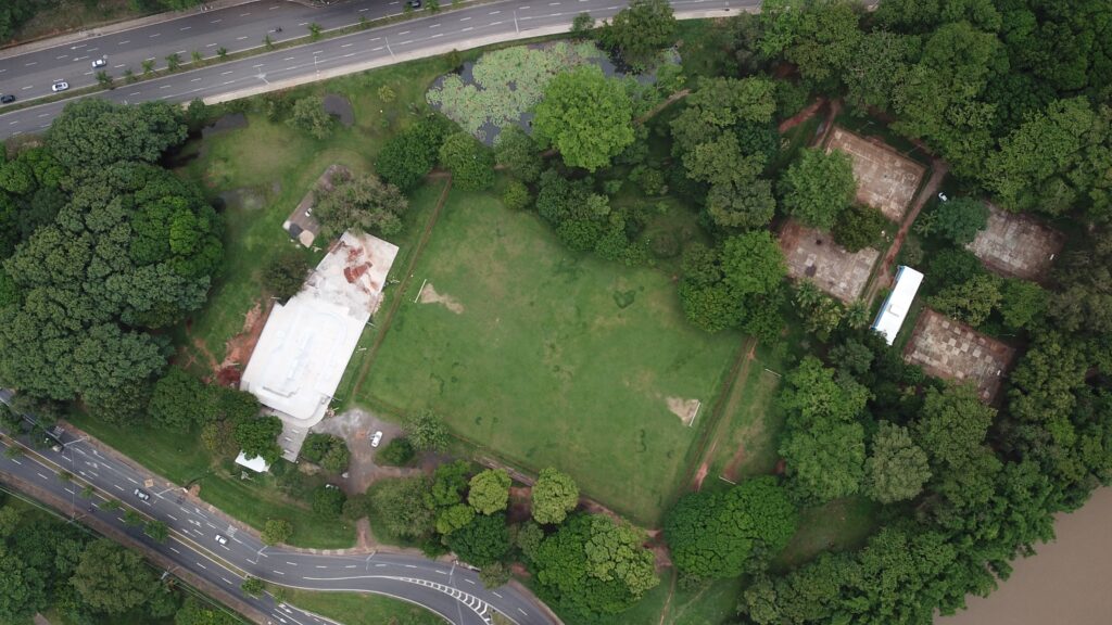 A imagem aérea revela uma área verde com diversas estruturas e elementos naturais. No centro, um campo de futebol gramado domina a paisagem, com traves em ambos os lados. À esquerda do campo, uma construção branca, possivelmente em reforma ou em construção, com áreas de concreto aparente e telhado branco. Ao redor do campo, a vegetação é abundante, com árvores frondosas e gramados. Um pequeno lago com plantas aquáticas, como lírios d'água, pode ser observado na parte superior esquerda da imagem, próximo a uma estrada com tráfego de veículos. Na parte direita da imagem, há um conjunto de quadras, possivelmente de tênis ou basquete, com pisos em tons de cinza e marcações visíveis. Uma construção longa e branca, semelhante a um contêiner, está posicionada entre algumas dessas quadras. A área é cercada por uma densa floresta, e um rio de águas escuras delimita a borda inferior direita da imagem. A presença de carros na estrada e em algumas áreas internas sugere que o local é acessível e possivelmente utilizado.
