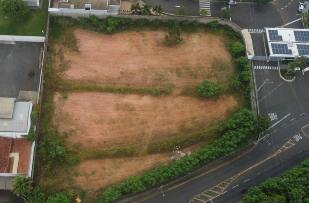 A imagem mostra uma vista aérea de um terreno baldio cercado por vegetação e edifícios. O terreno é predominantemente de terra vermelha, com algumas áreas de grama e arbustos dispersos. Ao redor do terreno, há uma rua asfaltada com sinalização de trânsito, incluindo faixas de pedestres e placas de "PARE". Em um dos lados, observa-se um edifício moderno com painéis solares no telhado, e em outro lado, há casas residenciais. A vegetação ao redor é densa, com árvores e arbustos que formam uma barreira natural entre o terreno e as áreas urbanizadas. A imagem parece ter sido capturada durante o dia, com iluminação natural.