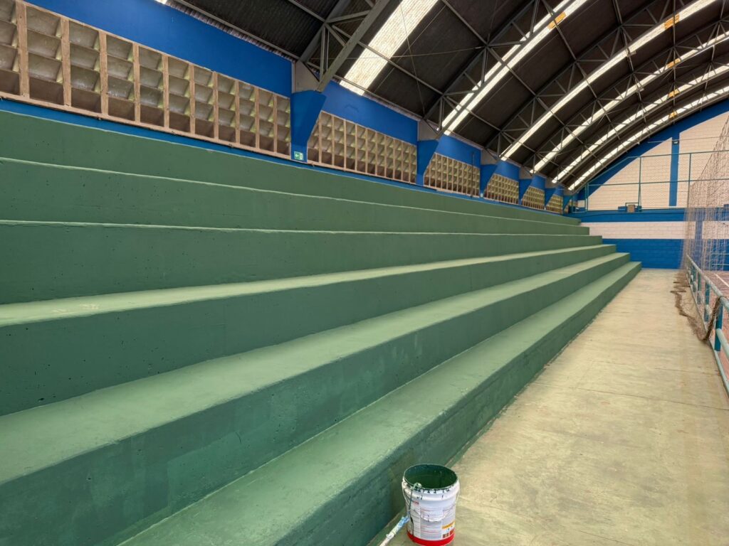 A imagem mostra arquibancadas de concreto pintadas de verde dentro de um ginásio coberto. As estruturas são dispostas em níveis e se estendem ao longo da lateral da quadra, oferecendo assentos para os espectadores. O espaço tem teto alto em arco com vigas metálicas e iluminação fluorescente, além de paredes azuis e brancas com blocos de ventilação na parte superior. No chão, próximo à base das arquibancadas, há um balde de tinta branco e vermelho com tampa verde, indicando manutenção ou pintura recente. A cena transmite a ideia de um ambiente esportivo em processo de conservação.
