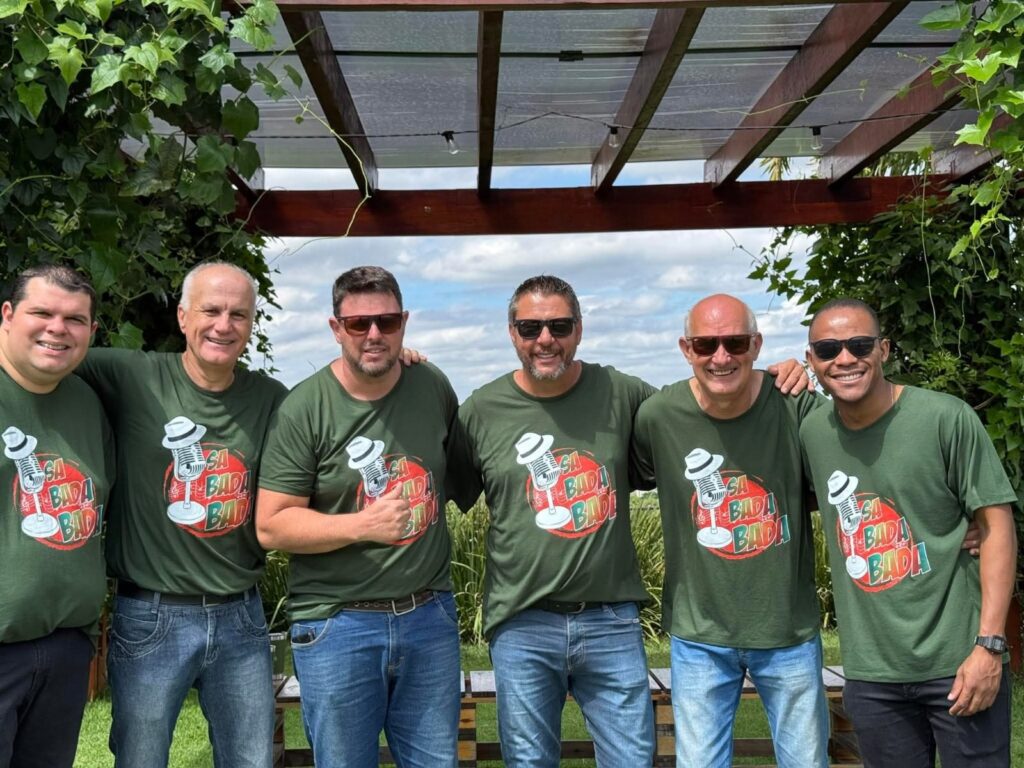 A imagem mostra um grupo de seis homens posando para uma foto ao ar livre. Todos estão vestindo camisetas verdes-escuras com um logo circular que apresenta um microfone estilizado com um chapéu e o texto "SA BADA BADA!". Eles estão sorrindo e parecem estar em um ambiente descontraído e amigável. O fundo inclui vegetação verde densa e uma estrutura de pergolado de madeira com cobertura translúcida. A iluminação é natural e sugere que a foto foi tirada durante o dia. Os homens estão organizados em duas fileiras, três na frente e três atrás, com os braços ao redor dos ombros uns dos outros, transmitindo um sentimento de camaradagem.
