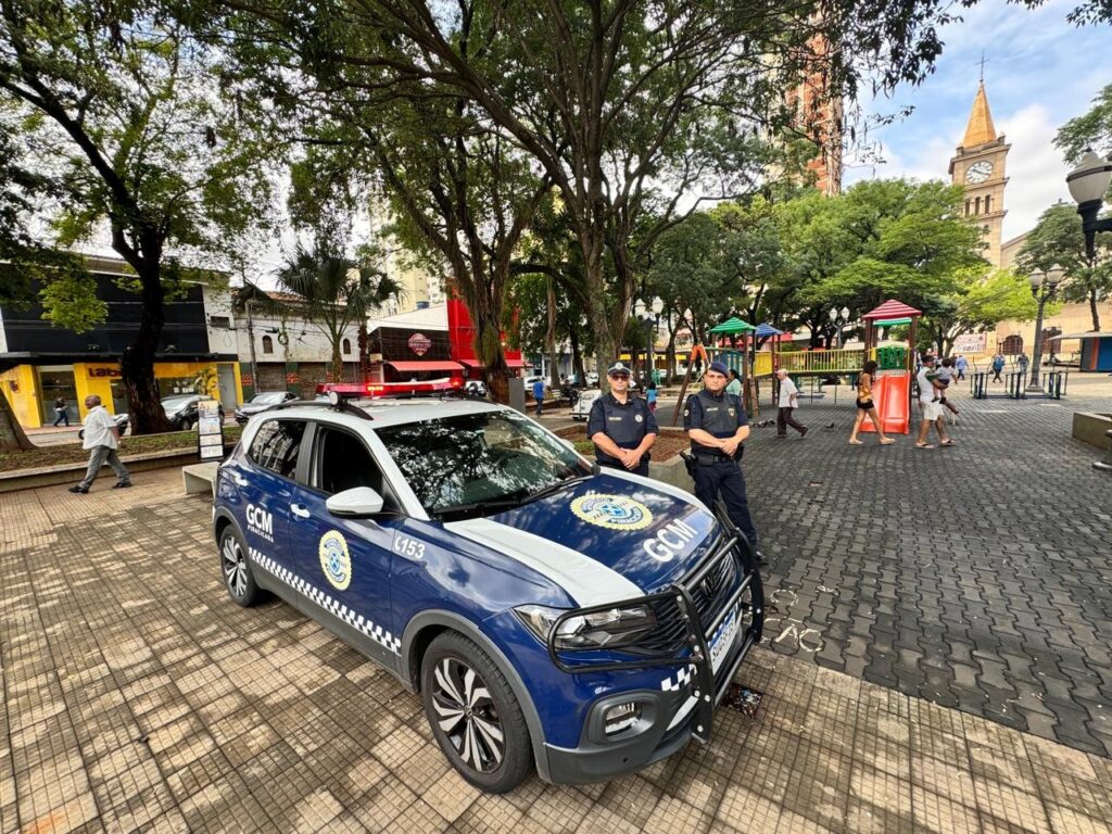 Na imagem, vê-se uma praça pública com um parque infantil ao fundo, onde pessoas circulam e aproveitam o espaço. Em primeiro plano, está estacionada uma viatura azul e branca da Guarda Civil Municipal (GCM), acompanhada por dois agentes uniformizados que permanecem ao lado do veículo. A cena transmite um ambiente de segurança e convivência comunitária, reforçado pela presença policial em um espaço de lazer.