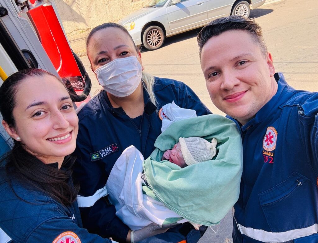 A imagem mostra três profissionais de saúde, dois homens e uma mulher, sorrindo para a câmera enquanto seguram um recém-nascido. O bebê, enrolado em uma manta verde e branca, usa um gorro de malha. Os profissionais usam uniformes azuis escuros com emblemas vermelhos e brancos. A mulher no centro está usando uma máscara facial branca. Ao fundo, um carro prateado pode ser visto. A cena parece ser um momento de alegria e celebração após o nascimento.