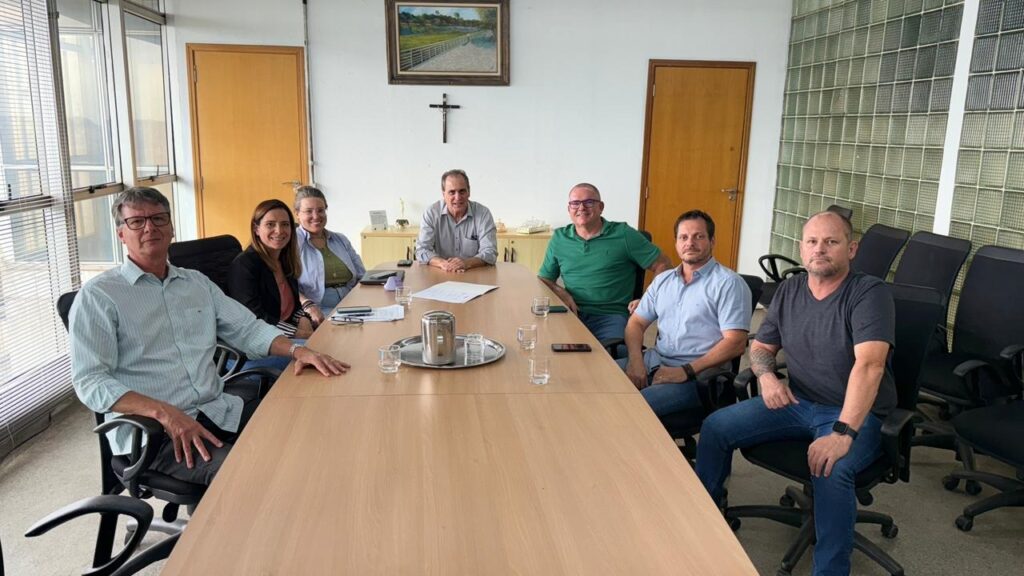 A imagem mostra um grupo de sete pessoas sentadas ao redor de uma mesa de madeira em uma sala de reuniões. Há uma grande janela com persianas do lado esquerdo da sala, uma porta de madeira à direita e uma parede com blocos de vidro translúcido no fundo. As pessoas estão vestidas casualmente. Do lado esquerdo, um homem de camisa listrada azul clara e óculos está sentado. Ao lado dele, uma mulher de jaqueta preta sobre uma blusa rosa e uma mulher de camisa verde clara e óculos. No centro da mesa, um homem de camisa cinza e um homem de camisa verde escura sorriem para a câmera. À direita, um homem de camisa azul clara e um homem de camisa grafite estão sentados. Na mesa, há copos com água, uma jarra de metal em uma bandeja e alguns papéis. Acima da mesa, há um quadro com uma pintura de uma paisagem e uma cruz.