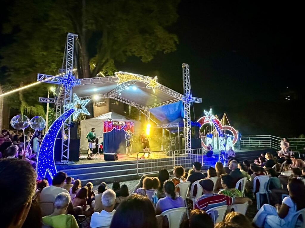 A imagem mostra uma cena noturna ao ar livre, com uma multidão reunida para assistir a uma apresentação em um palco iluminado. O palco está decorado com luzes azuis e estrelas amarelas, e há um letreiro com "RISO SHOW" escrito. No palco, alguns artistas estão se apresentando, com um músico tocando bateria e outros em pé. A plateia, composta por muitas pessoas sentadas em cadeiras e em pé, observa a performance atentamente. Ao redor do palco, há decorações luminosas em forma de arco e estrelas, criando uma atmosfera festiva. Ao fundo, árvores escuras e o céu noturno completam a cena.