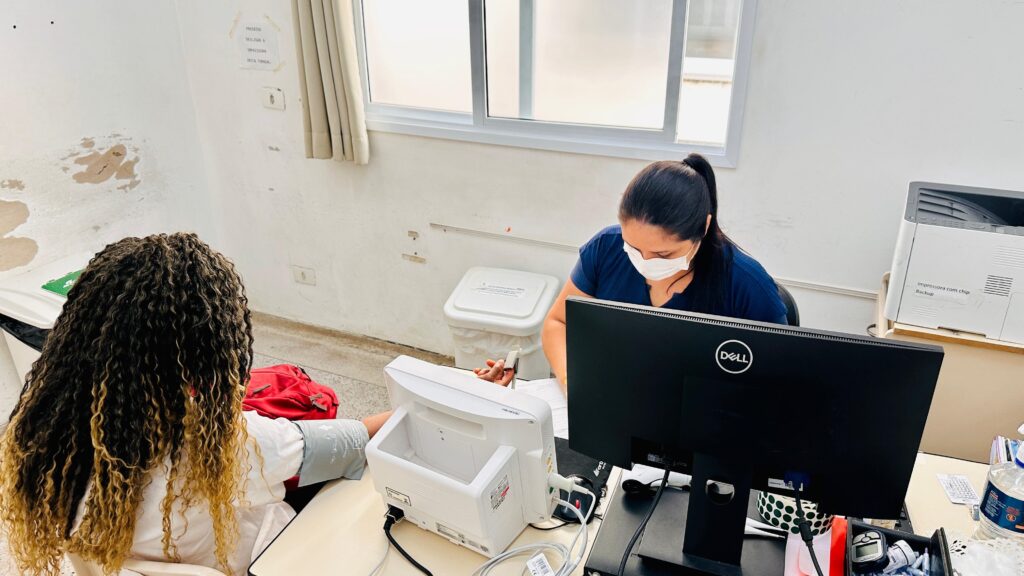 A imagem mostra duas mulheres em um ambiente que parece ser uma clínica ou posto de saúde. Uma delas, com longos cabelos cacheados e tingidos de loiro, está sentada de costas para a câmera, usando uma camisa branca e uma braçadeira de aparelho médico. A outra mulher, atrás de uma mesa com um computador Dell e uma impressora, usa uma máscara facial branca e uma blusa azul. Há uma lixeira branca ao fundo, perto de uma janela com cortinas. A parede ao lado da primeira mulher apresenta sinais de umidade e descascamento da pintura.