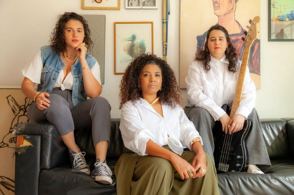 A imagem mostra três mulheres sentadas em um sofá de couro preto, com um fundo composto por várias obras de arte emolduradas. A mulher da esquerda, com cabelos castanhos ondulados, veste um colete jeans sobre uma blusa branca aberta e calças cinzas. Ela usa joias douradas, incluindo um colar de pérolas e pulseiras. No centro, uma mulher com cabelos cacheados escuros usa uma camisa branca e calças verdes. Ela também está adornada com joias douradas, como um colar e anéis. A mulher da direita, com cabelos escuros e lisos, veste uma camisa branca e calças cinzas. Ela está segurando um baixo elétrico preto, com a mão apoiada nas cordas. Todas as mulheres parecem estar olhando para a câmera com expressões sérias. O ambiente sugere um espaço criativo ou de ensaio.