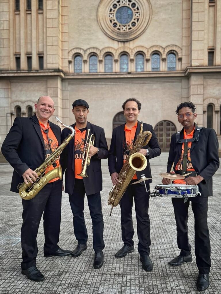 A imagem mostra quatro homens posando para uma foto em frente a um edifício com arquitetura clássica. Todos os homens estão vestindo ternos pretos sobre camisas laranja vibrantes. Da esquerda para a direita: * O primeiro homem está sorrindo e segurando um saxofone dourado. * O segundo homem, usando um boné preto, segura uma trombeta dourada. * O terceiro homem está sorrindo e segurando um saxofone dourado maior. * O quarto homem, usando óculos, está posicionado atrás de uma caixa de bateria azul, segurando baquetas. O edifício atrás deles tem uma fachada de pedra com arcos e uma grande janela circular ornamentada no topo. O chão em primeiro plano é pavimentado com um padrão quadriculado. A iluminação sugere que a foto foi tirada durante o dia, possivelmente ao entardecer, dada a luz suave.