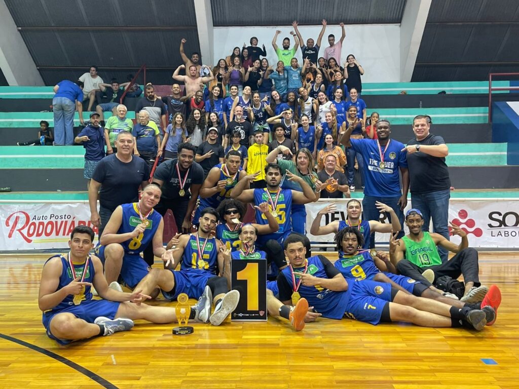 A imagem mostra uma equipe de basquete comemorando uma vitória em uma quadra. Os jogadores, vestindo uniformes azuis e amarelos, posam para a foto com medalhas no pescoço e alguns segurando troféus. Um grande número "1" está em destaque no centro da imagem, simbolizando o primeiro lugar. A equipe parece feliz e orgulhosa, com alguns membros fazendo gestos de vitória. Ao fundo, espectadores e outros membros da equipe estão sentados nas arquibancadas, também celebrando. A atmosfera geral é de alegria e triunfo