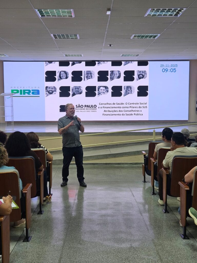 A imagem mostra uma sala de conferências com um palestrante em pé, segurando um microfone e falando para uma audiência sentada em cadeiras de madeira. Atrás do palestrante, há um grande telão exibindo o logotipo da "PREFEITURA PIRA" à esquerda, e uma série de fotos de pessoas, juntamente com o título "Conselhos de Saúde: O Controle Social e o Financiamento como Pilares do SUS - Atribuições dos Conselheiros x Financiamento da Saúde Pública". O telão também exibe a data "29-11-2025" e a hora "09:05". O palestrante, um homem de meia-idade com barba e óculos, veste uma camisa de manga curta e calças escuras. A audiência, composta por homens e mulheres, está sentada em fileiras de cadeiras de madeira, voltada para o palestrante. O ambiente parece ser uma instituição pública, possivelmente uma prefeitura ou um centro de convenções.