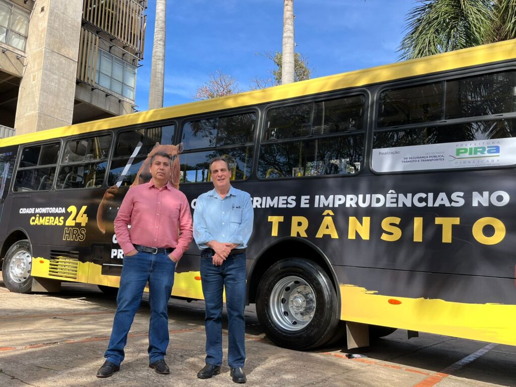 Na imagem, dois homens estão posicionados diante de um ônibus com design preto e amarelo, que exibe mensagens de segurança pública em destaque. O veículo traz inscrições como “Cidade monitorada câmeras 24 hrs” e “Crimes e imprudências no trânsito”, indicando uma campanha municipal voltada à vigilância urbana e ao combate a comportamentos perigosos no tráfego. Ao fundo, aparecem um prédio moderno de concreto e vidro, palmeiras e um céu azul, compondo um cenário urbano organizado.