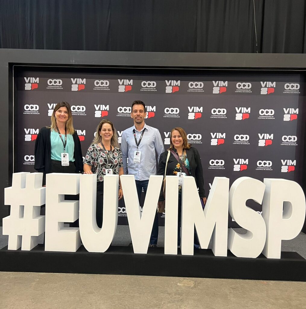 A imagem mostra quatro pessoas em pé atrás de letras brancas grandes que formam a frase "#EUVIMSP". As pessoas parecem estar em um evento ou conferência, pois há um banner de fundo com logotipos repetidos. O banner tem o texto "VIM CCD COORDENADORIA DE CONTROLE DE DOENÇAS" repetido várias vezes. As quatro pessoas estão sorrindo e olhando para a câmera. Elas estão vestindo crachás, sugerindo que são participantes ou expositores do evento. As letras grandes "#EUVIMSP" são o ponto focal da imagem, com as pessoas posicionadas atrás delas. O fundo é escuro, o que faz com que as letras brancas se destaquem. A iluminação é clara, iluminando as pessoas e as letras.