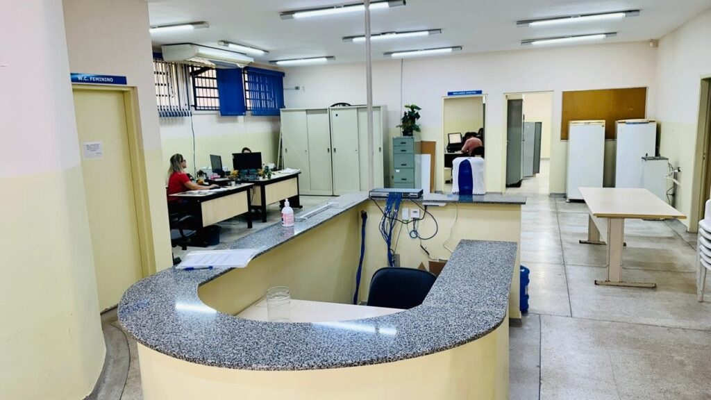 A imagem mostra o interior de um escritório ou recepção, com uma bancada em destaque no primeiro plano. A bancada tem um acabamento em granito escuro e curvo, e há alguns itens sobre ela, como papéis e um copo. Atrás da bancada, vemos um ambiente de trabalho com mesas, computadores e armários. Há uma mulher sentada em uma das mesas, trabalhando em um computador. Ao fundo, há portas que levam a outras salas, e em uma delas, também há uma pessoa trabalhando. Há também refrigeradores e uma mesa comprida no fundo do escritório. A iluminação é feita por lâmpadas fluorescentes no teto. A porta à esquerda indica "W.C. FEMININO".