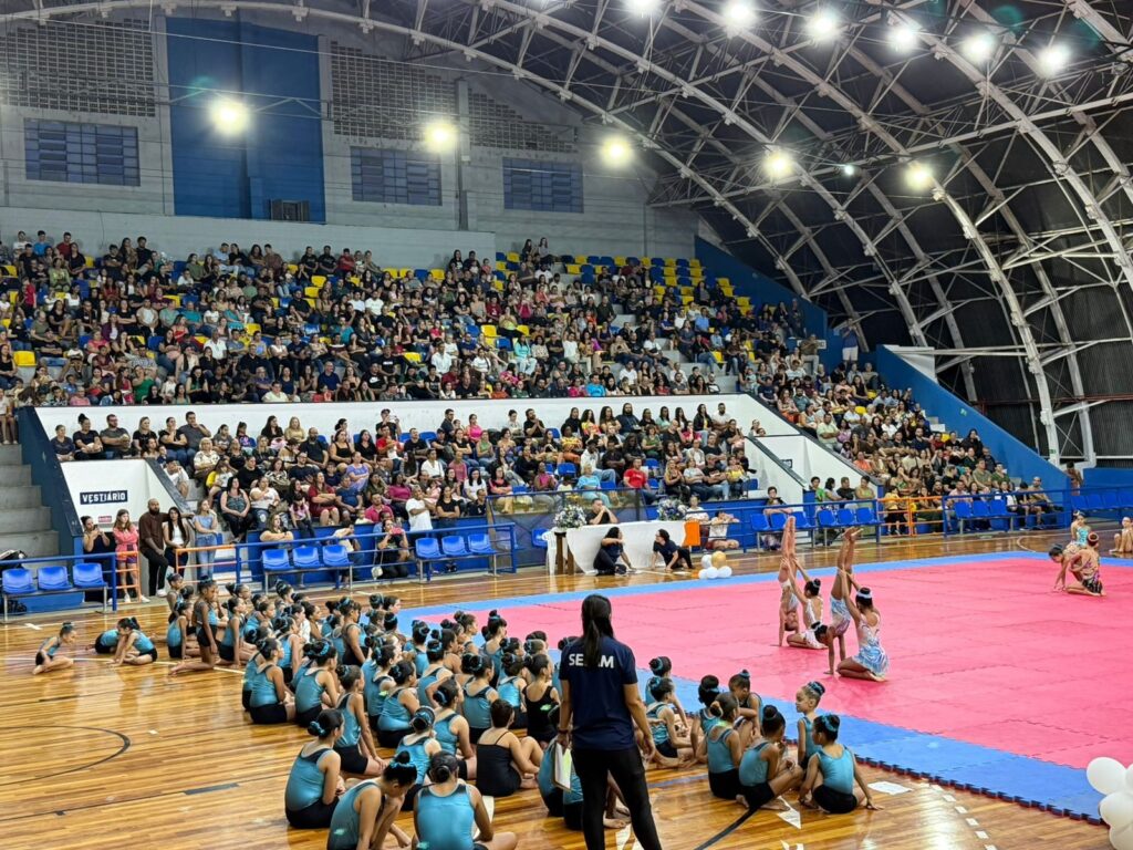 A imagem retrata um evento de ginástica rítmica em um ginásio esportivo. Ao fundo, arquibancadas coloridas estão repletas de espectadores, observando atentamente a competição. No centro da quadra, sobre um tapete rosa e azul, jovens ginastas demonstram suas habilidades em apresentações individuais e em grupo. Algumas ginastas estão em posições acrobáticas, enquanto outras aguardam sua vez, vestindo trajes esportivos. Uma instrutora, com uma camiseta escrito "SE M", observa as participantes. O ambiente é de celebração e esporte, com a iluminação do ginásio destacando as cores e os movimentos.