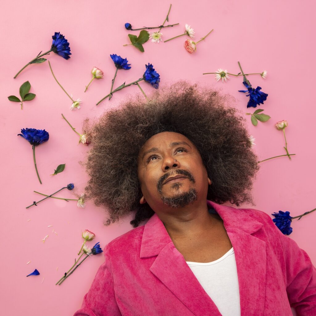 A imagem mostra uma pessoa deitada em um fundo rosa vibrante, com a cabeça e parte do corpo visíveis. A pessoa tem um cabelo afro volumoso e escuro, e usa uma jaqueta rosa choque sobre uma camiseta branca. Ao redor da pessoa, espalhadas pelo fundo, há várias flores azuis e brancas, algumas com pequenos botões de rosa. A iluminação é suave e direcionada, criando um ambiente lúdico e artístico. A pessoa olha para cima, com uma expressão contemplativa.