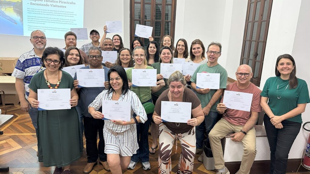 A imagem mostra um grupo de pessoas, a maioria mulheres, reunidas em uma sala, segurando certificados. Elas parecem ter acabado de concluir um curso ou workshop, pois todas sorriem e seguram seus certificados com orgulho. No fundo, há um projetor com slides que parecem ser sobre turismo em Piracicaba, com o título "Receptivo Turístico Piracicaba – Encantando Visitantes". A sala parece ser um local de aprendizado, possivelmente uma sala de aula ou auditório, com janelas altas e uma porta de madeira ao fundo. As pessoas estão vestindo roupas casuais e diversas, e a atmosfera geral é de celebração e realização. A maioria dos certificados exibem o logo do SENAC, indicando que o curso foi oferecido por essa instituição.