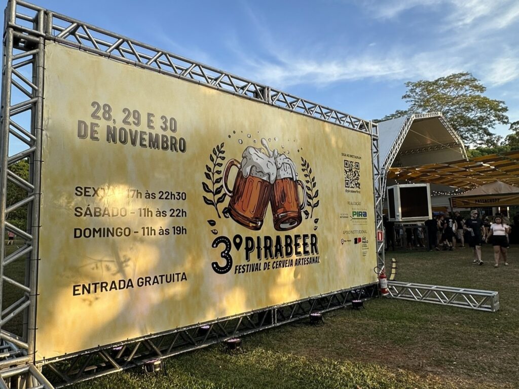 A imagem mostra um outdoor de um festival de cerveja artesanal chamado "3º Pirabeer". O evento ocorrerá nos dias 28, 29 e 30 de novembro, com horários variados para cada dia: sexta-feira das 17h às 22h30, sábado das 11h às 22h e domingo das 11h às 19h. A entrada é gratuita. Há também informações sobre a realização do evento e apoio institucional, além de um QR code e o @pirabeerfest para seguir no Instagram. Ao fundo, observa-se uma estrutura de palco e tendas, indicando que o evento está em andamento ou prestes a começar.