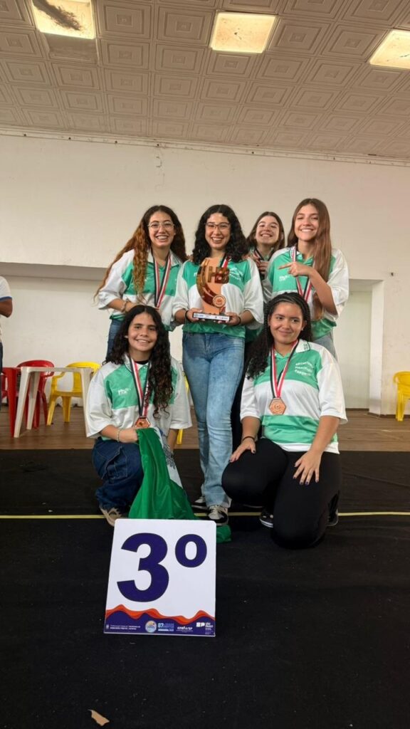 A imagem mostra um grupo de seis jovens mulheres sorrindo, posando para uma foto. Elas parecem estar em um evento de premiação, pois a maioria usa medalhas e uma delas segura um troféu. Há uma placa no chão com o número "3º", indicando que elas conquistaram o terceiro lugar. As jovens vestem uniformes brancos com detalhes em verde e têm expressões alegres e orgulhosas. Algumas estão em pé, enquanto outras estão ajoelhadas ou sentadas em uma plataforma. Ao fundo, vemos cadeiras coloridas e um teto com luminárias. A atmosfera geral é de celebração e conquista.