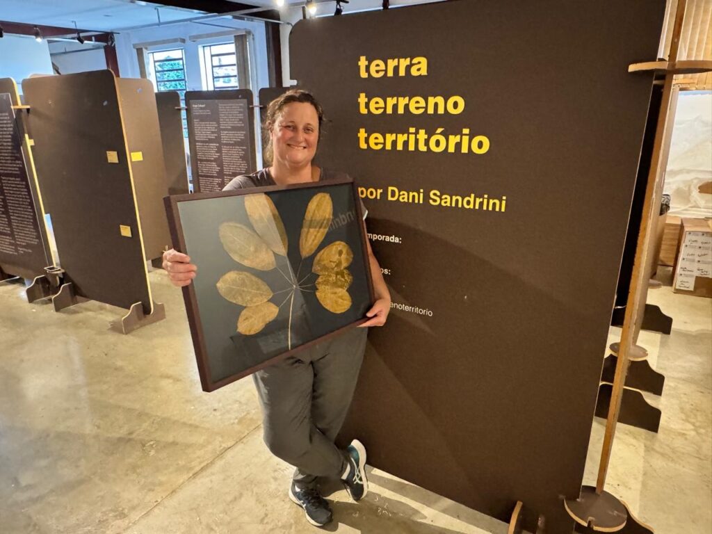 A mulher sorri para a câmera enquanto segura uma moldura com folhas secas. Ela está em pé em frente a um grande painel marrom com texto amarelo escrito nele. Há também uma exposição de arte com mais painéis ao redor dela. O chão é de concreto e a iluminação parece ser artificial.