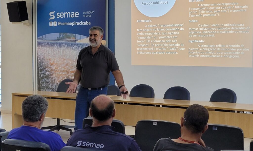 A imagem mostra uma sala de aula ou auditório com um homem em pé, apresentando algo para um grupo de pessoas sentadas. O homem, que parece ser o palestrante, está sorrindo e gesticulando com as mãos. Ele veste uma camisa polo escura e calça jeans. Ao fundo, há um banner com o logotipo "semae" e a frase "UM NOVO TEMPO", além de um @seguidor com o nome "@semaepiracicaba". Há também uma projeção na parede com slides de uma apresentação. Um dos slides explica a etimologia da palavra "responsabilidade", detalhando sua origem no latim, a raiz e o sufixo, e o significado da palavra. As pessoas sentadas na plateia estão de costas para a câmera, e algumas delas usam camisetas com o logotipo "semae". A iluminação da sala parece ser artificial, vinda de luminárias no teto.