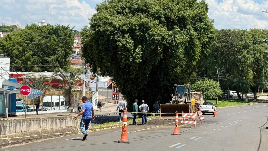 Na imagem, observa-se uma cena de manutenção viária em área urbana. Alguns trabalhadores, vestidos com camisas azuis e calças jeans, atuam no local delimitado por cones laranja e fitas de isolamento, indicando uma zona de obras. Dentro da área restrita, há um veículo de construção pesado, semelhante a uma pá carregadeira, além de uma caminhonete branca estacionada próxima.