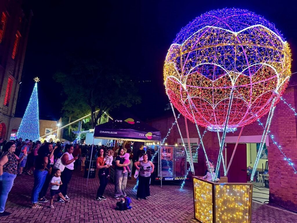 A imagem retrata uma cena noturna vibrante e festiva, dominada por uma grande instalação de luzes em formato de balão. Este balão, suspenso no ar, é adornado com uma profusão de luzes em tons de azul, vermelho e amarelo, criando um espetáculo visual impressionante. Ao fundo, uma árvore de Natal iluminada com luzes azuis se destaca, complementando o ambiente festivo. Uma multidão de pessoas, incluindo famílias com crianças, está reunida, observando as atrações e aproveitando a atmosfera. Há também uma barraca com o logo "RVATELECOM" e um veículo com a inscrição "MEU AMIGO BEER", sugerindo um evento com patrocinadores e opções de bebidas. A iluminação geral da cena, com luzes cintilantes e coloridas, cria um clima mágico e acolhedor.