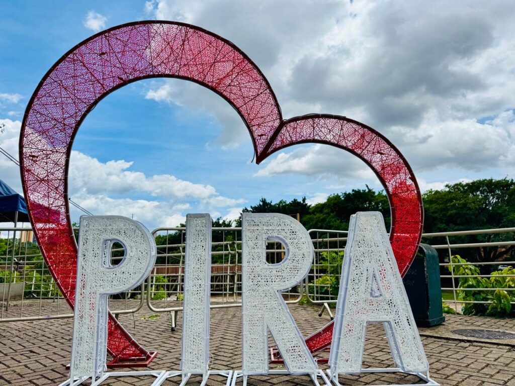 A imagem mostra uma grande instalação artística ao ar livre com as letras "PIRA" em destaque, feitas de um material branco que parece ser luzes de LED ou um material translúcido similar. As letras são grandes e tridimensionais, com uma estrutura que sugere que elas podem ser iluminadas. Atrás das letras, há um grande coração vermelho translúcido, também feito de um material que parece ser uma rede ou malha fina, com um padrão intrincado. O coração parece estar emoldurando uma parte do céu. O fundo da imagem é um céu azul com nuvens brancas fofas. Ao fundo, à direita, há uma linha de árvores verdes densas e, à esquerda, uma estrutura azul que pode ser parte de uma tenda ou edifício. Há também uma cerca metálica baixa e um cesto de lixo verde, sugerindo que a instalação está em uma área pública, como um parque ou praça. O chão é pavimentado com blocos de pedra em um padrão de espinha de peixe. A iluminação sugere que é um dia claro e ensolarado.