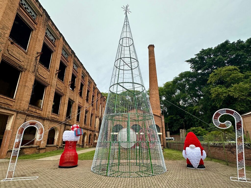 A imagem exibe uma cena festiva de Natal em frente a um edifício histórico de tijolos, que parece abandonado ou em ruínas, com janelas quebradas e sem vidros. No centro, destaca-se uma grande árvore de Natal em formato cônico, feita de uma estrutura metálica verde e adornada com inúmeras luzes brancas que formam um efeito cintilante. A árvore é ladeada por decorações natalinas. À esquerda, há um grande enfeite em formato de bengala de doce, feito de luzes brancas. Ao lado da bengala, encontra-se uma bota vermelha gigante, decorada com luzes, que simula uma bota do Papai Noel, com uma gola branca e caixas de presente empilhadas no topo. À direita da árvore, há uma figura de um gnomo natalino, também feito de luzes, com um chapéu vermelho pontudo e uma barba branca volumosa. Mais à direita, outro enfeite de bengala de doce, semelhante ao da esquerda, completa a composição. Ao fundo, observa-se uma chaminé de tijolos, característica de antigas construções industriais, e árvores frondosas que contrastam com a arquitetura rústica do edifício. O céu está nublado, conferindo uma atmosfera um pouco sombria ao cenário, mas as luzes natalinas criam um ponto de alegria e celebração.